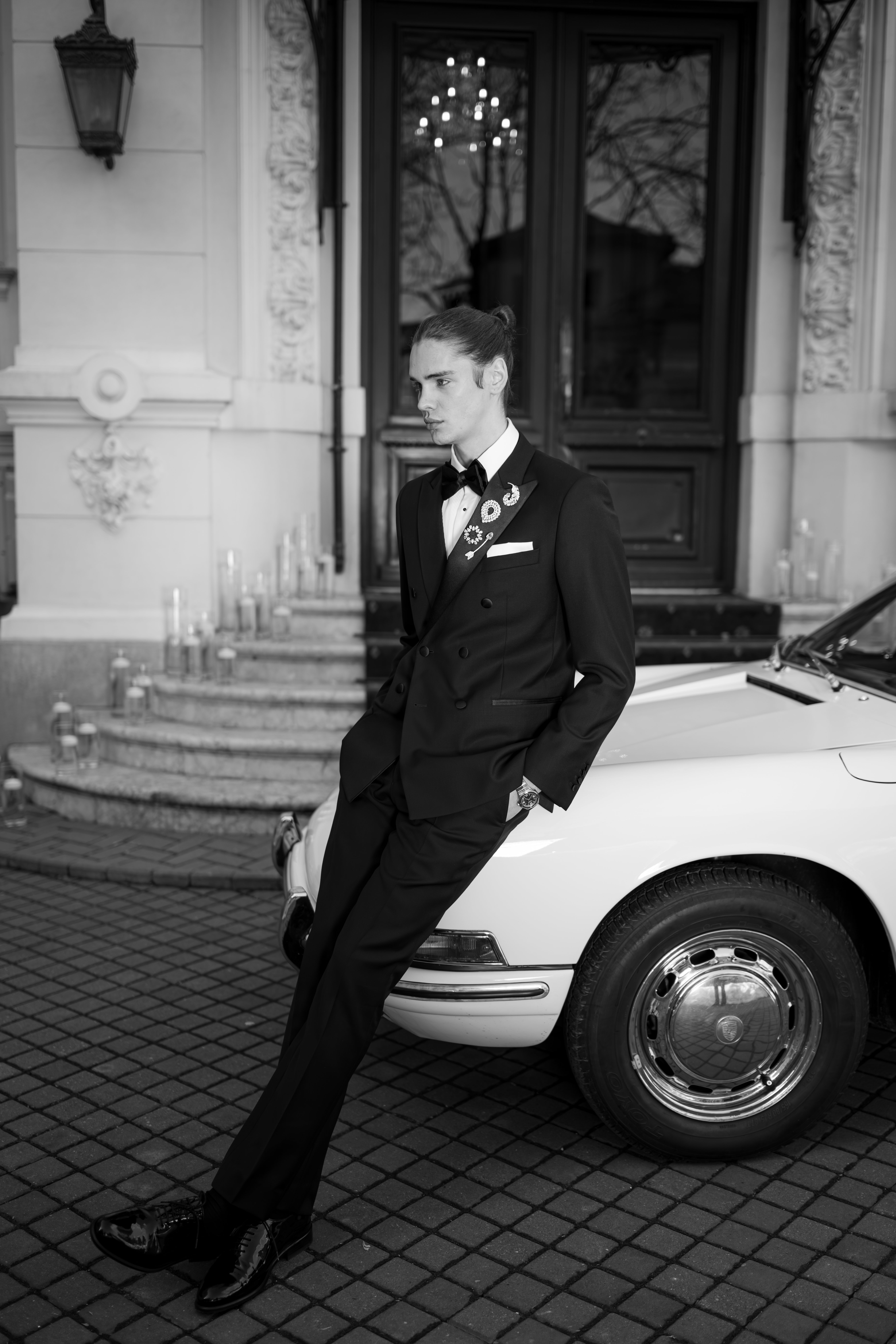 Mire în tuxedo negru lângă Porsche alb vintage în fața Palatului Noblesse — editorial alb-negru nuntă 2026