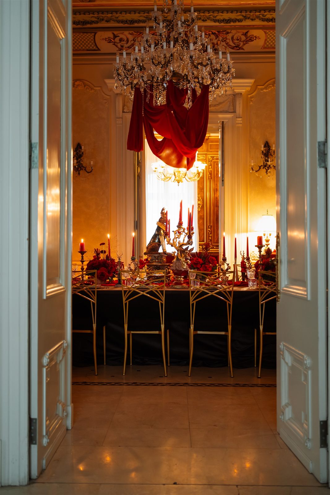 Perspectivă prin uși către salonul de gală cu candelabru de cristal și masă festivă în roșu la Palatul Noblesse