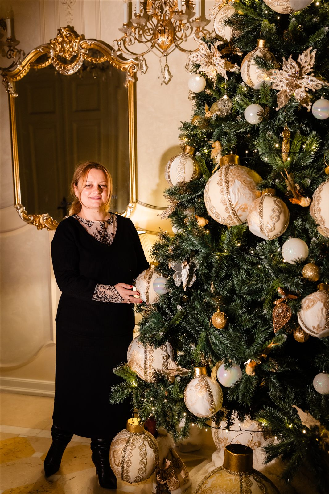 Gazda Palatului Noblesse lângă bradul de Crăciun cu globuri albe și aurii, sub candelabru