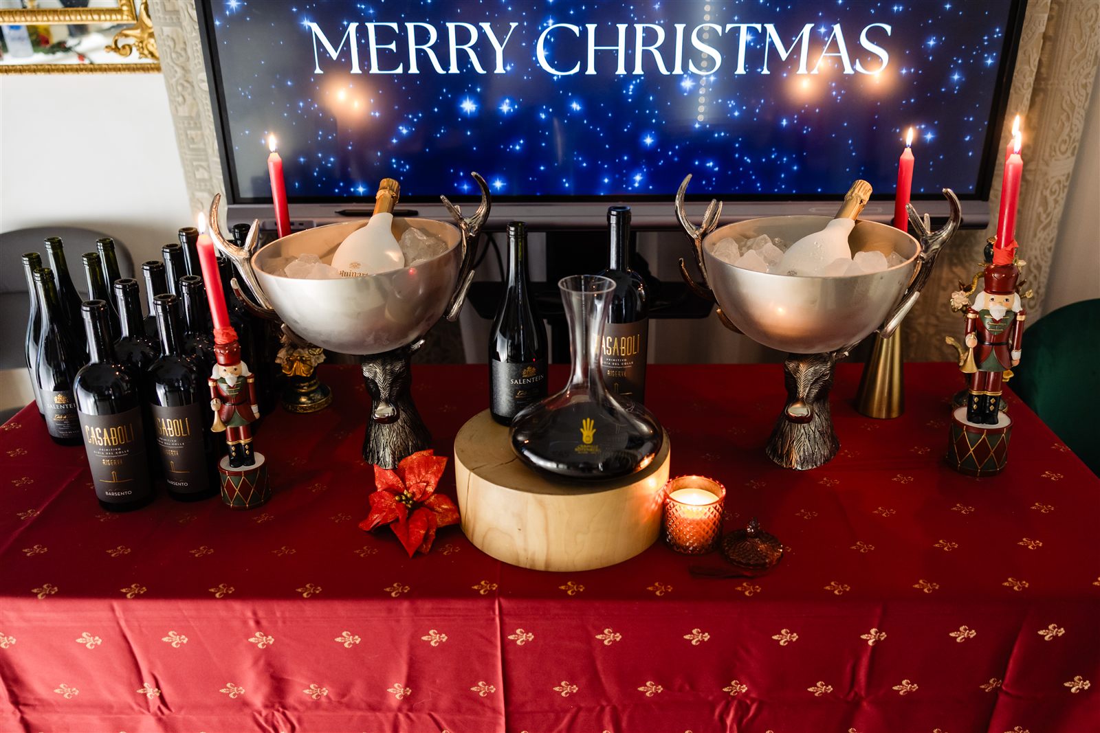 Masă cu vinuri și șampanie în frapiere, mesaj Merry Christmas pe ecran