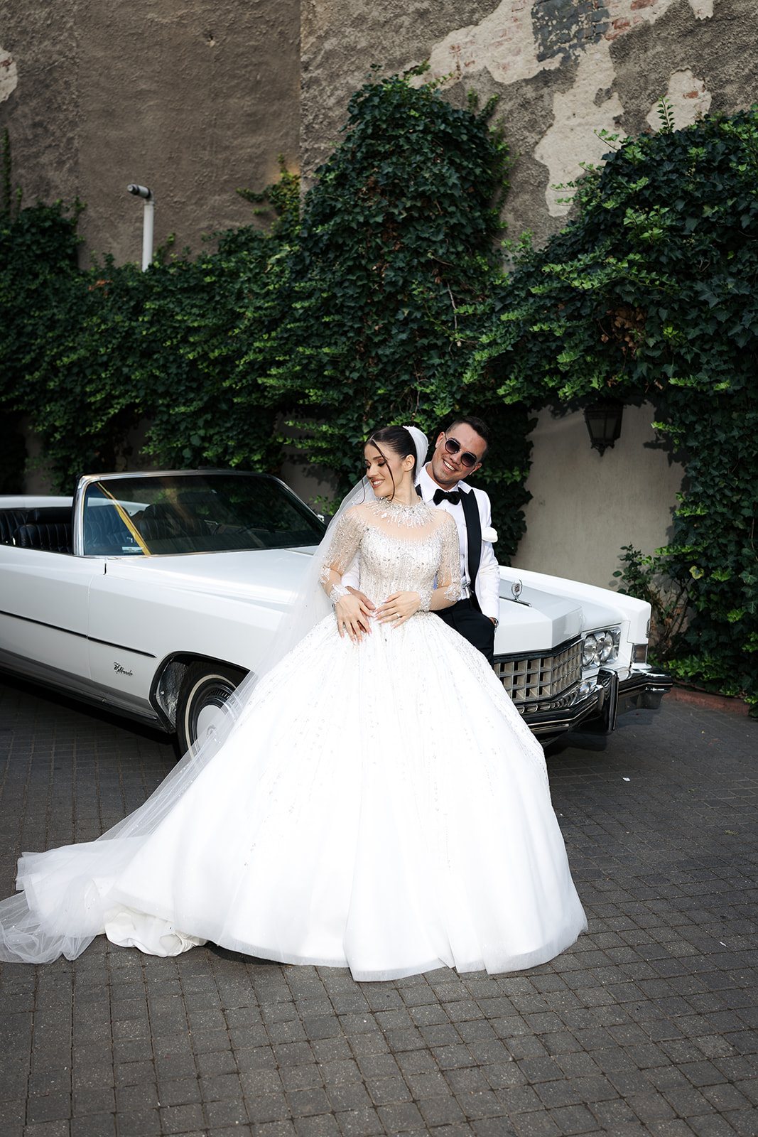 Mireasă în rochie cu trenă lungă lângă Cadillac Eldorado vintage, curtea Palatului Noblesse